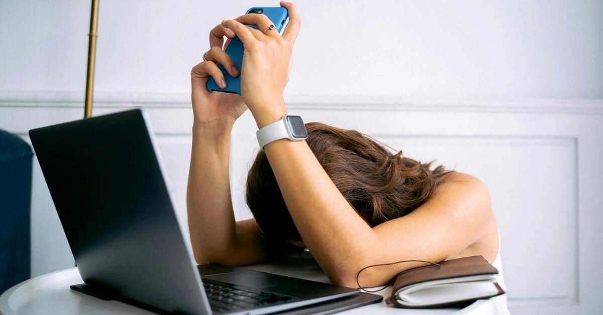 Woman sitting at desk, frustrated with her head on the laptop, holding a phone in her hands