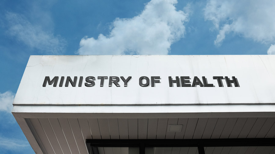A low-angle shot of a white building facade featuring the words "MINISTRY OF HEALTH" in bold, black, 3D block letters. The background shows a bright blue sky with soft, scattered white clouds.