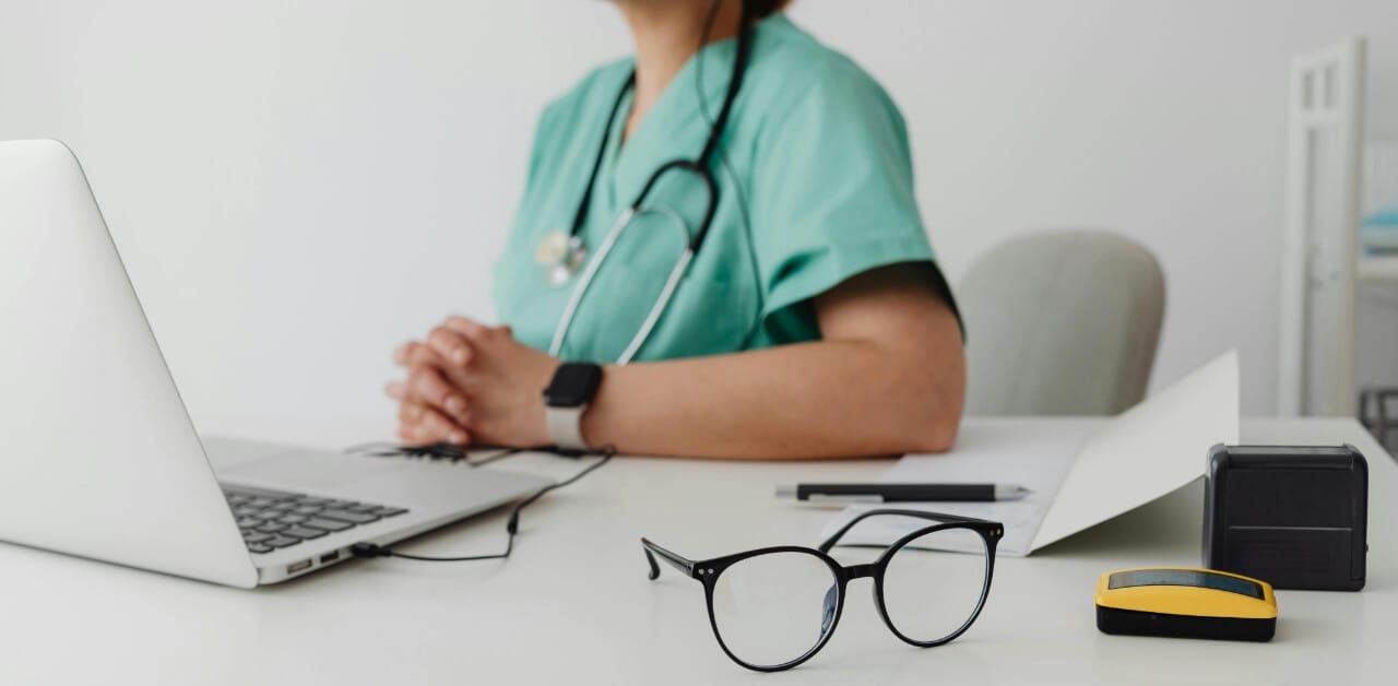 Nurse sitting behind laptop, with a pair of glasses on the desk.